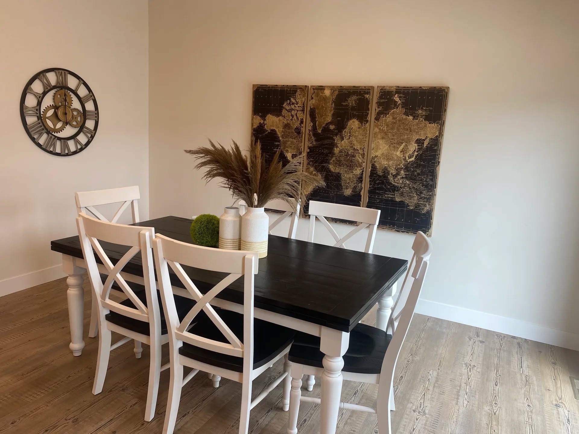 A dining area with a dark wooden table, white chairs, and a centerpiece of vases and grass. A world map art piece and a clock adorn the walls.