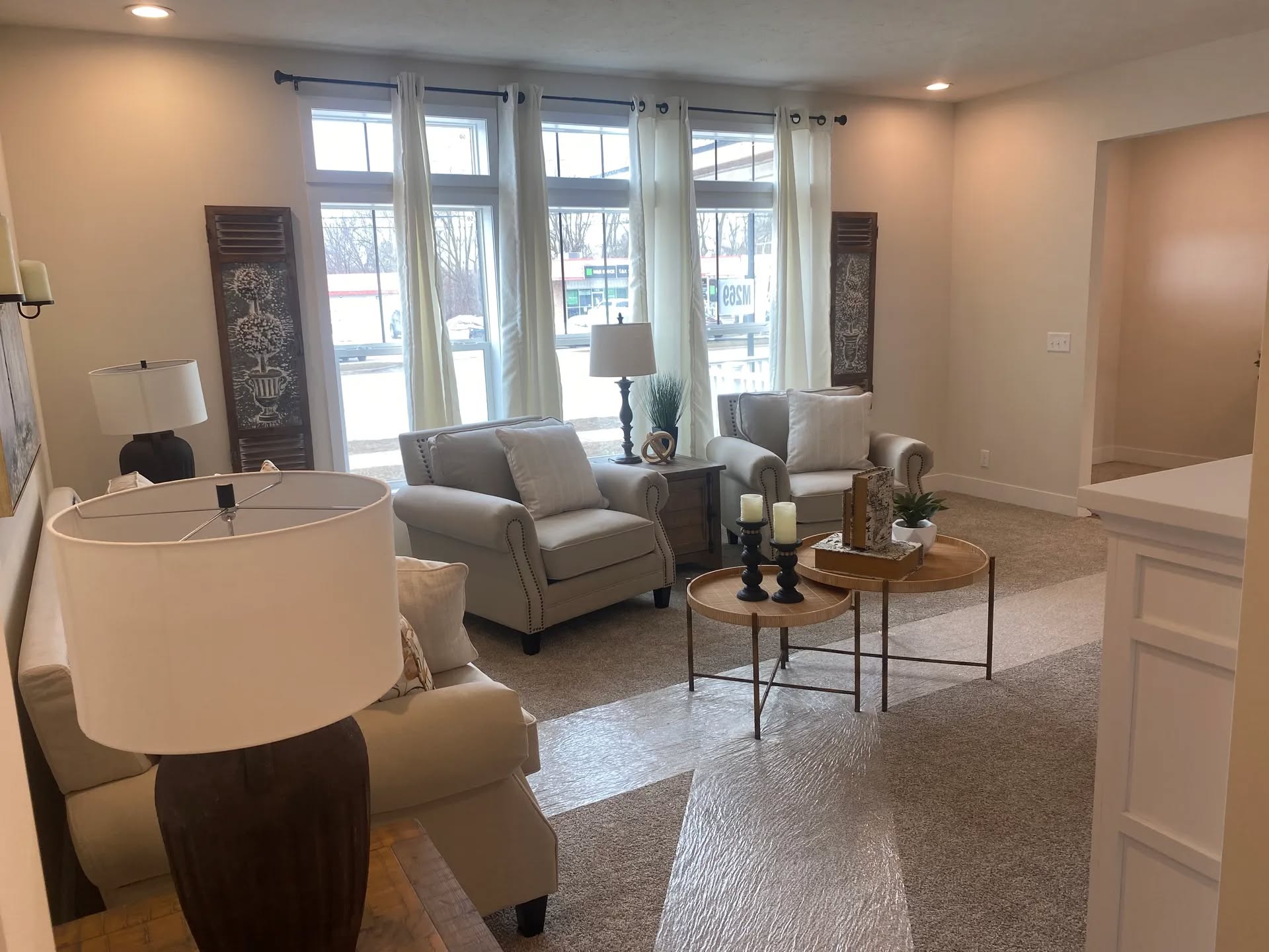 Cozy living room with beige sofas, a round wooden coffee table, and floor-to-ceiling windows with sheer curtains. Warm lighting creates a welcoming atmosphere.