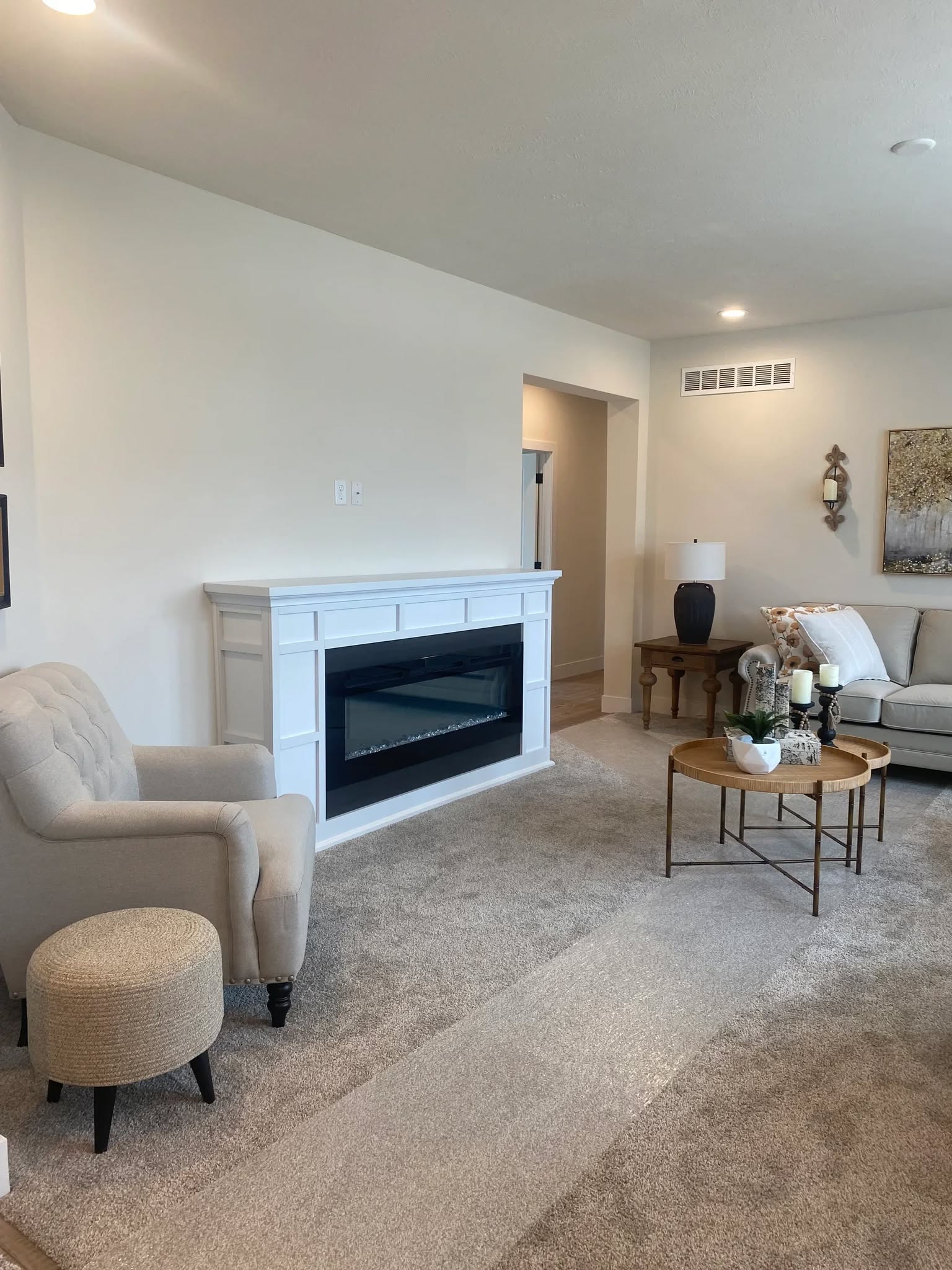 Cozy living room with beige walls and carpet, featuring a white fireplace, a plush armchair with ottoman, and a round coffee table with decor items.