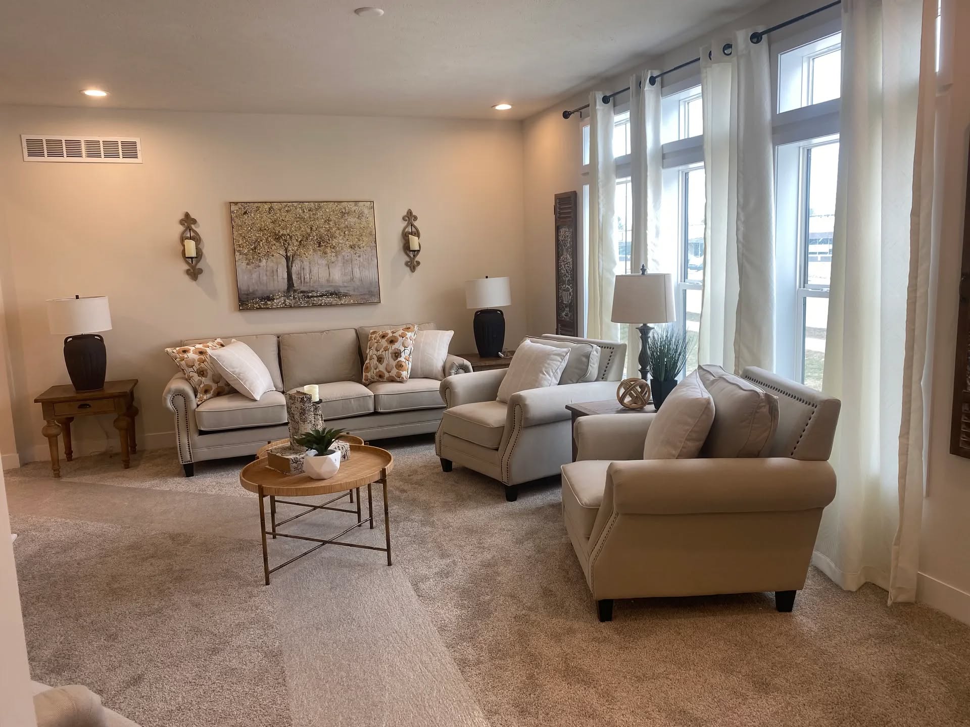 A cozy living room with beige sofas and armchairs arranged around a circular wooden coffee table. Large windows with sheer curtains let in natural light.