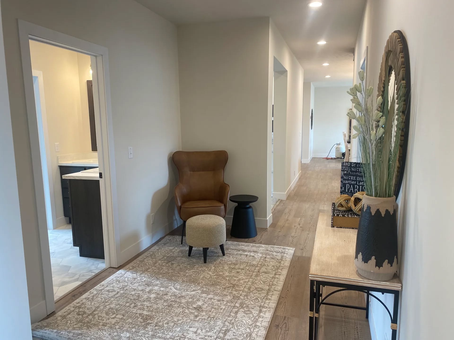Sleek hallway with a brown armchair and ottoman on a patterned rug. Open door reveals a bathroom. Vase with plants and decor on a console table. Warm and inviting ambiance.
