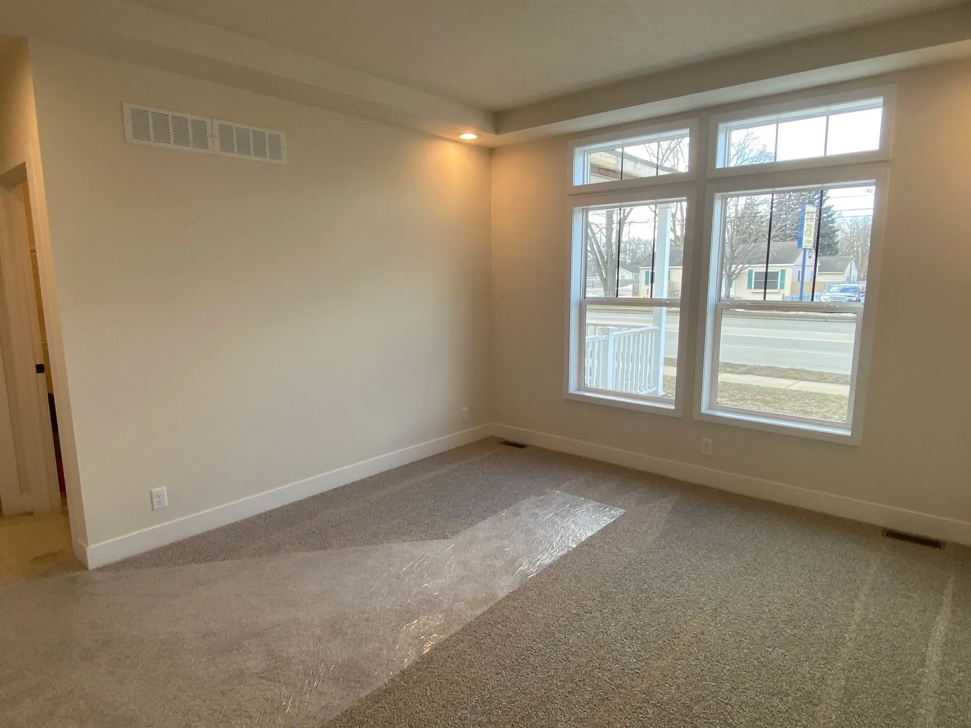Bright, empty room with beige walls and carpet, large window overlooking a street and houses. Natural light creates a calm, inviting atmosphere.