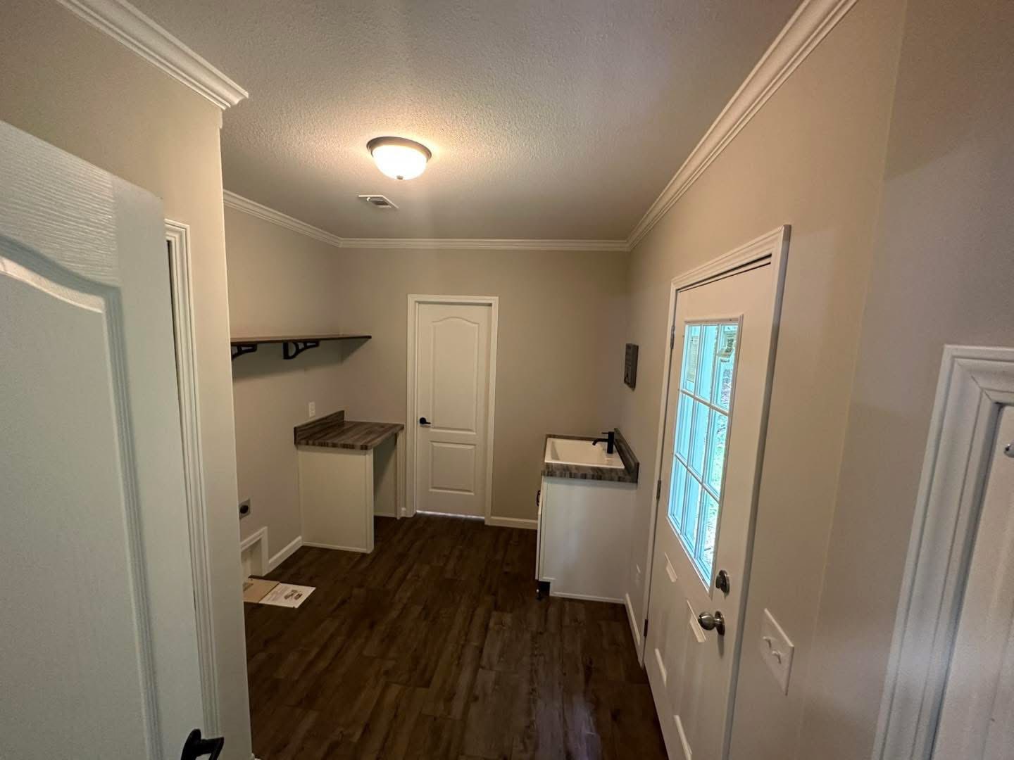 Bright utility room with wood flooring, white walls, and a ceiling light. Features include a countertop, open shelving, a door with window panes, and a sink.