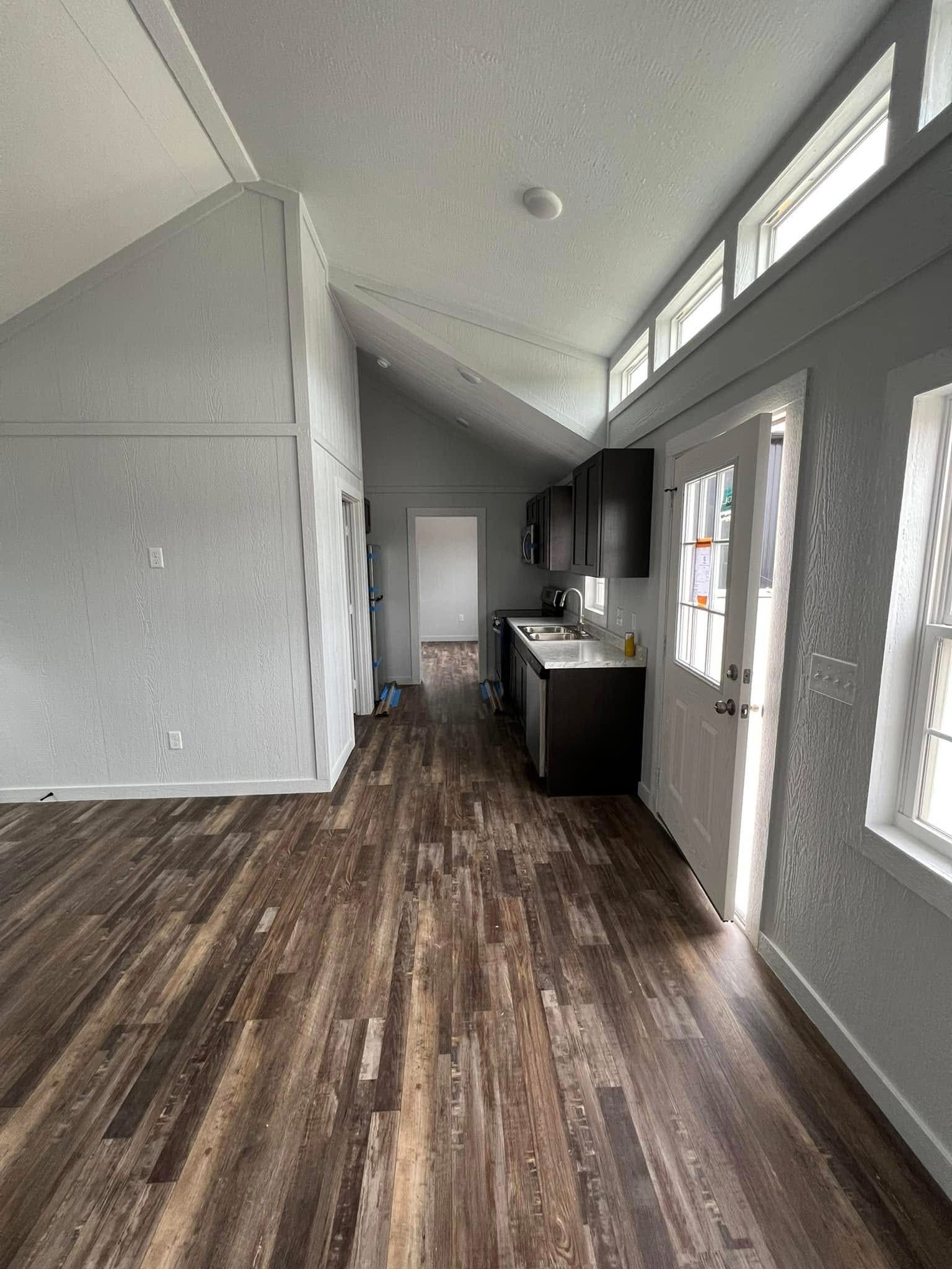 Modern kitchen in a small home with wood flooring, dark cabinets, and sleek counters. Bright natural light from high windows creates an airy feel.