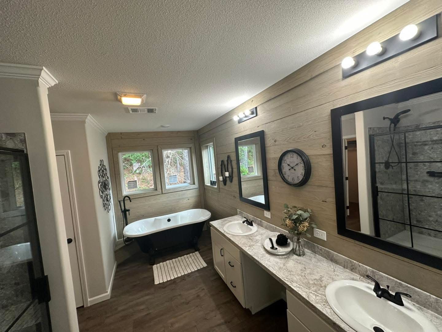 Modern bathroom with a black freestanding tub by large windows, dual sinks on a marble countertop, wood-patterned walls, mirrors, and a wall clock. Bright and elegant.