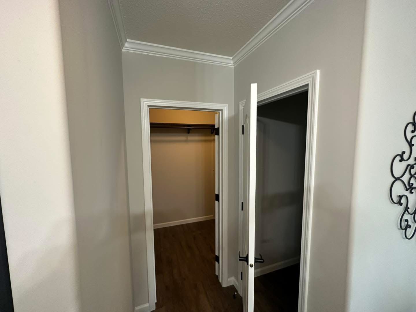 A small, empty closet with a white door is open, revealing a wooden floor. The closet is part of a hallway with light gray walls and white trim.