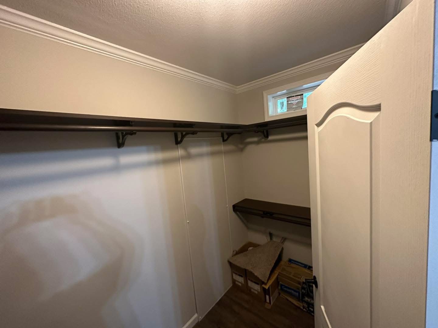 Walk-in closet with beige walls and dark wooden shelves. A small window provides light. Floor has cardboard boxes. The space feels organized and minimalistic.