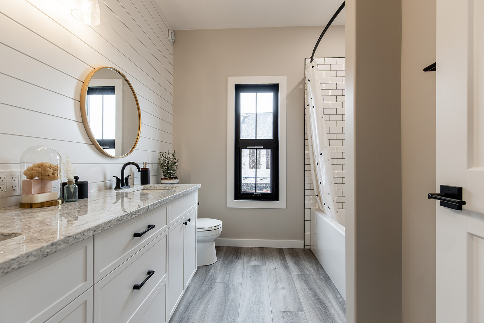 A modern bathroom with white shiplap walls and a light wood floor. Features include a round mirror, a granite countertop, and a bathtub with a subway tile surround. The atmosphere is clean and bright.