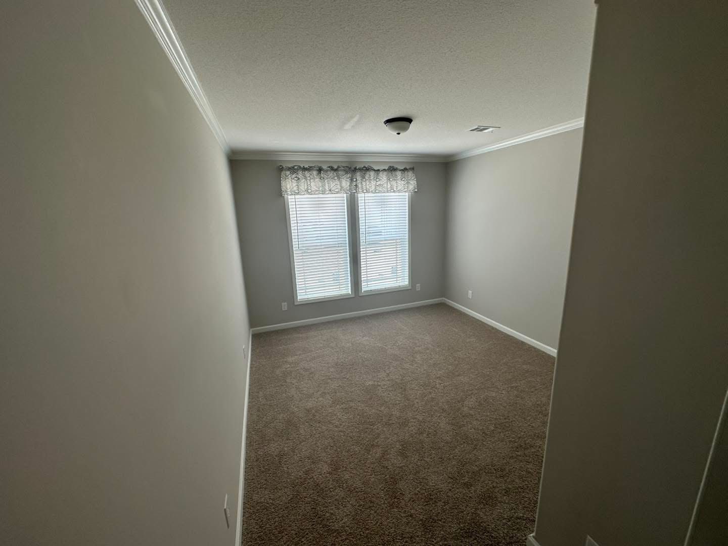 A small, empty room with beige carpet and light gray walls, featuring two closed, shuttered windows with a decorative valance, conveying a calm, neutral tone.