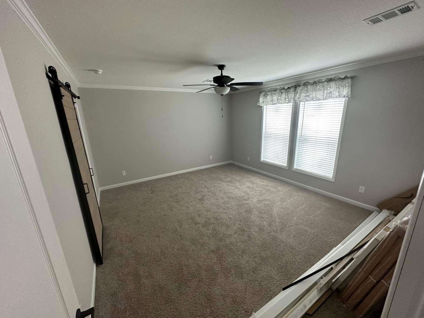 An empty room with grey walls and carpet, featuring large windows with floral valances, a ceiling fan, and a sliding barn door. Wooden planks rest on the floor.