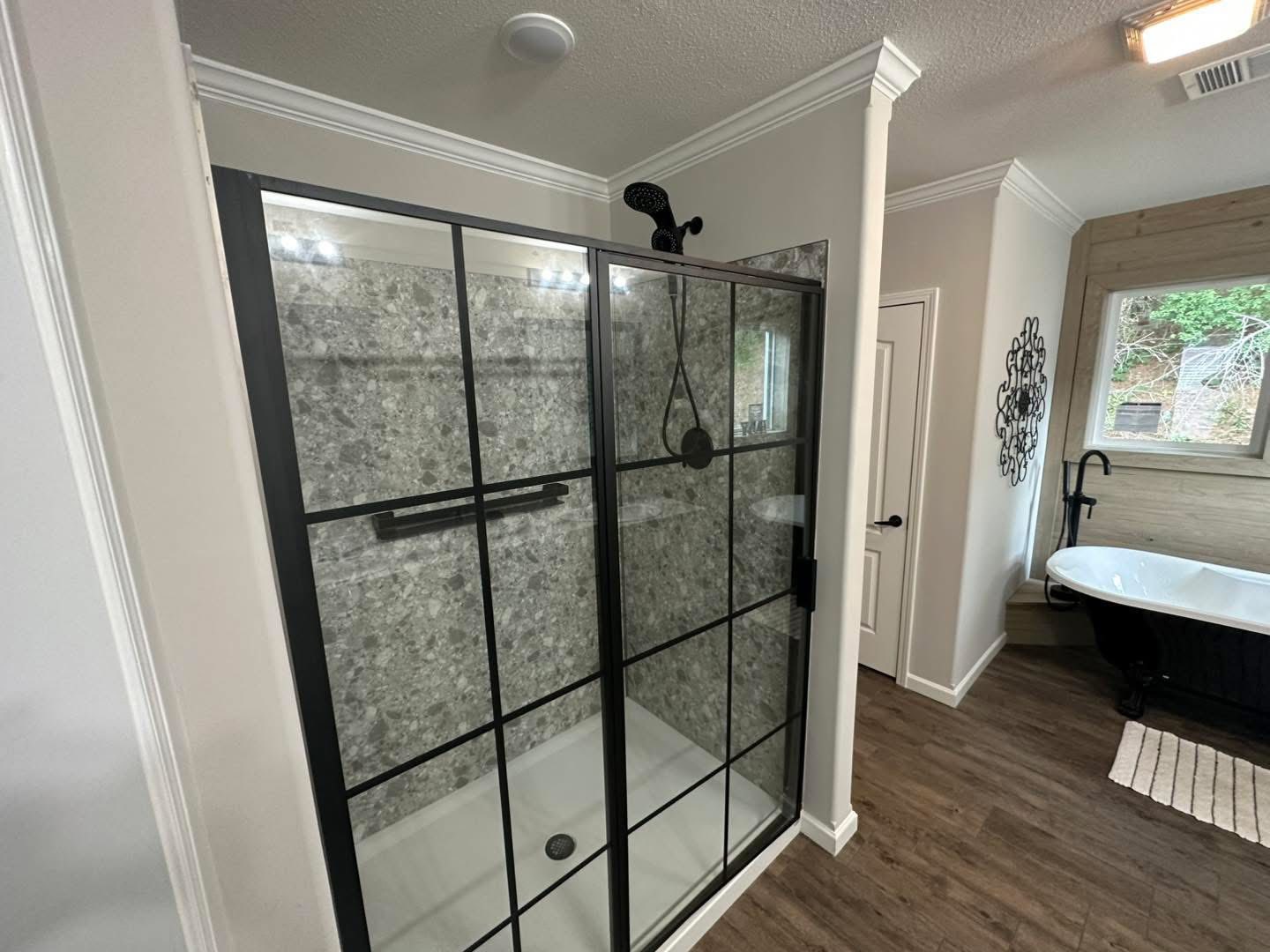 Modern bathroom with a glass shower featuring black grid framing and gray stone walls. Adjacent is a white freestanding tub and a large window.