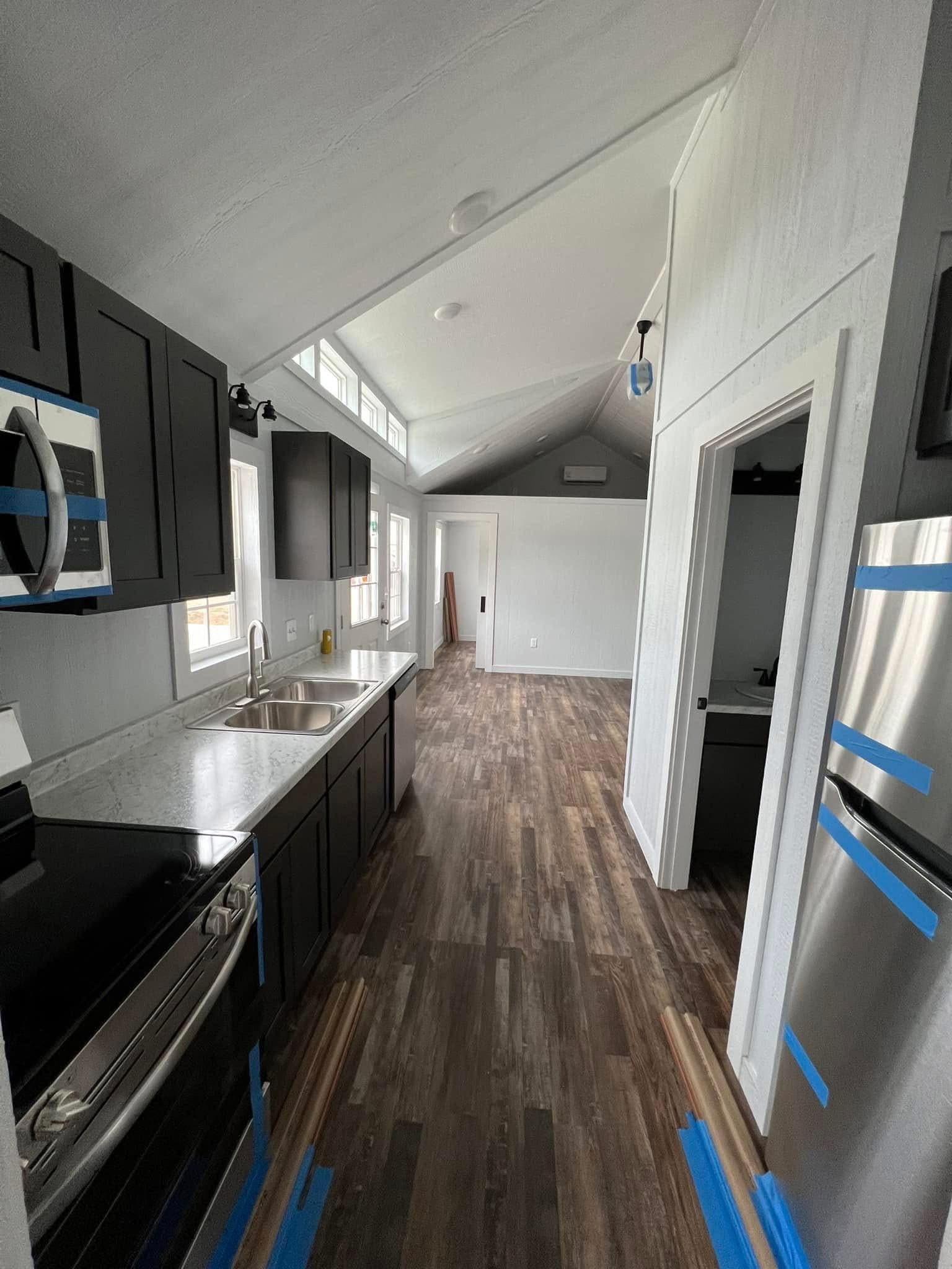 Modern, narrow kitchen with dark cabinets, stainless steel appliances, and a white countertop. Sunlight streams through adjacent windows, highlighting the wood floor, creating a bright and airy atmosphere.
