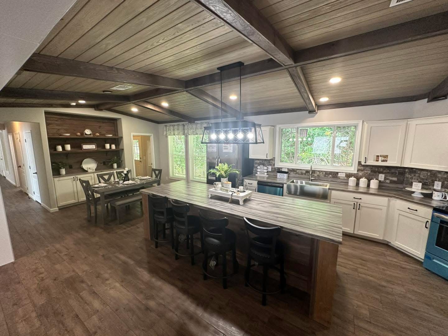 A cozy kitchen with a rustic wooden ceiling and floor. Features a long wooden island with stools and a modern light fixture. Bright windows create a welcoming atmosphere.