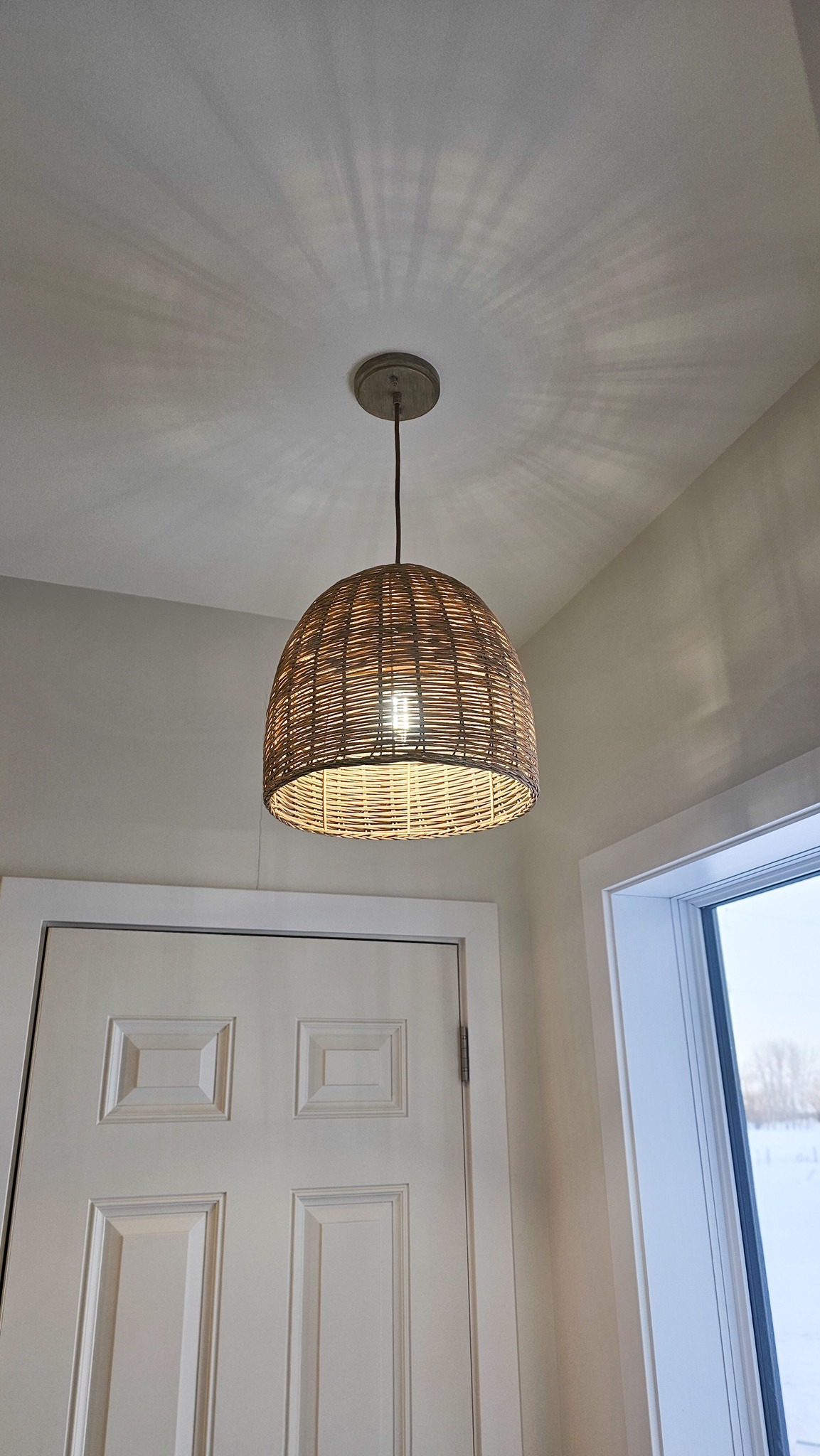 A woven wicker lampshade hangs from the ceiling, casting intricate shadow patterns. It illuminates a bright room with a white door and window, evoking a cozy atmosphere.