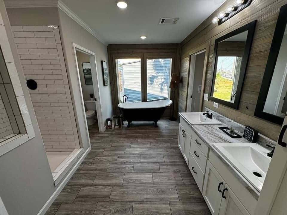 A modern bathroom with wooden floors, featuring a freestanding black bathtub by large windows, a double vanity with mirrors, and a tiled shower.