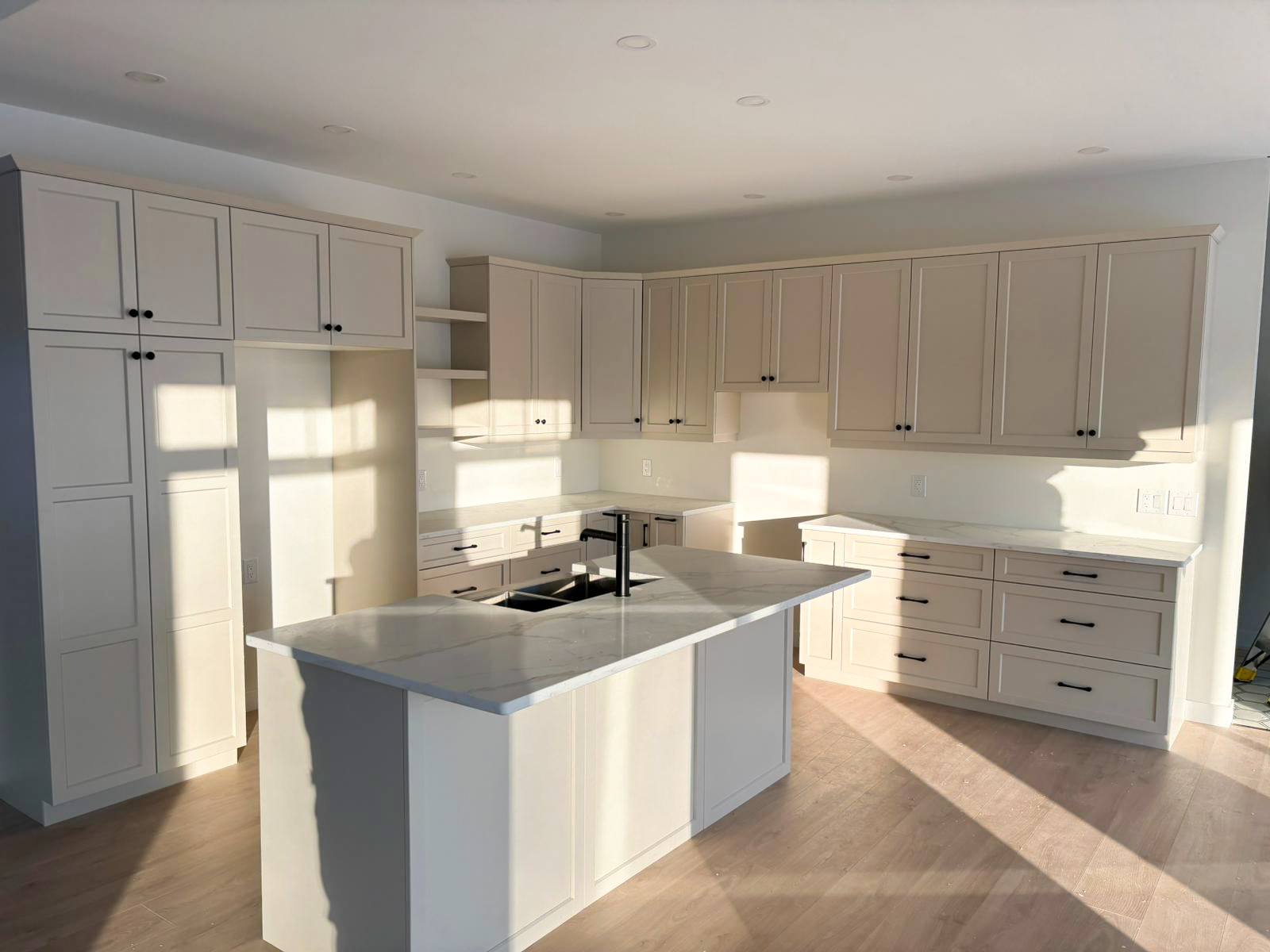 Modern kitchen with white cabinets, a marble island, and black hardware. Sunlight casts shadows on the wooden floor, creating a warm, inviting atmosphere.