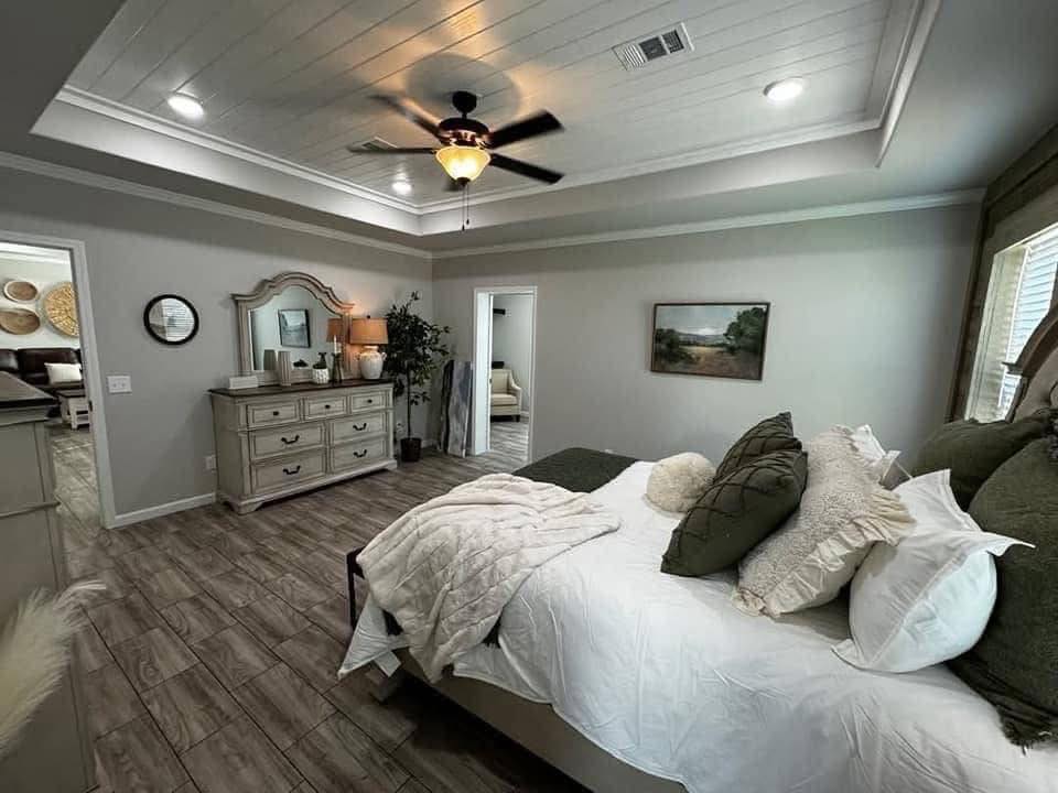 Cozy bedroom with wood flooring, tray ceiling, and ceiling fan. A neatly made bed with green and white pillows faces a dresser with a large mirror. Tranquil and inviting atmosphere.