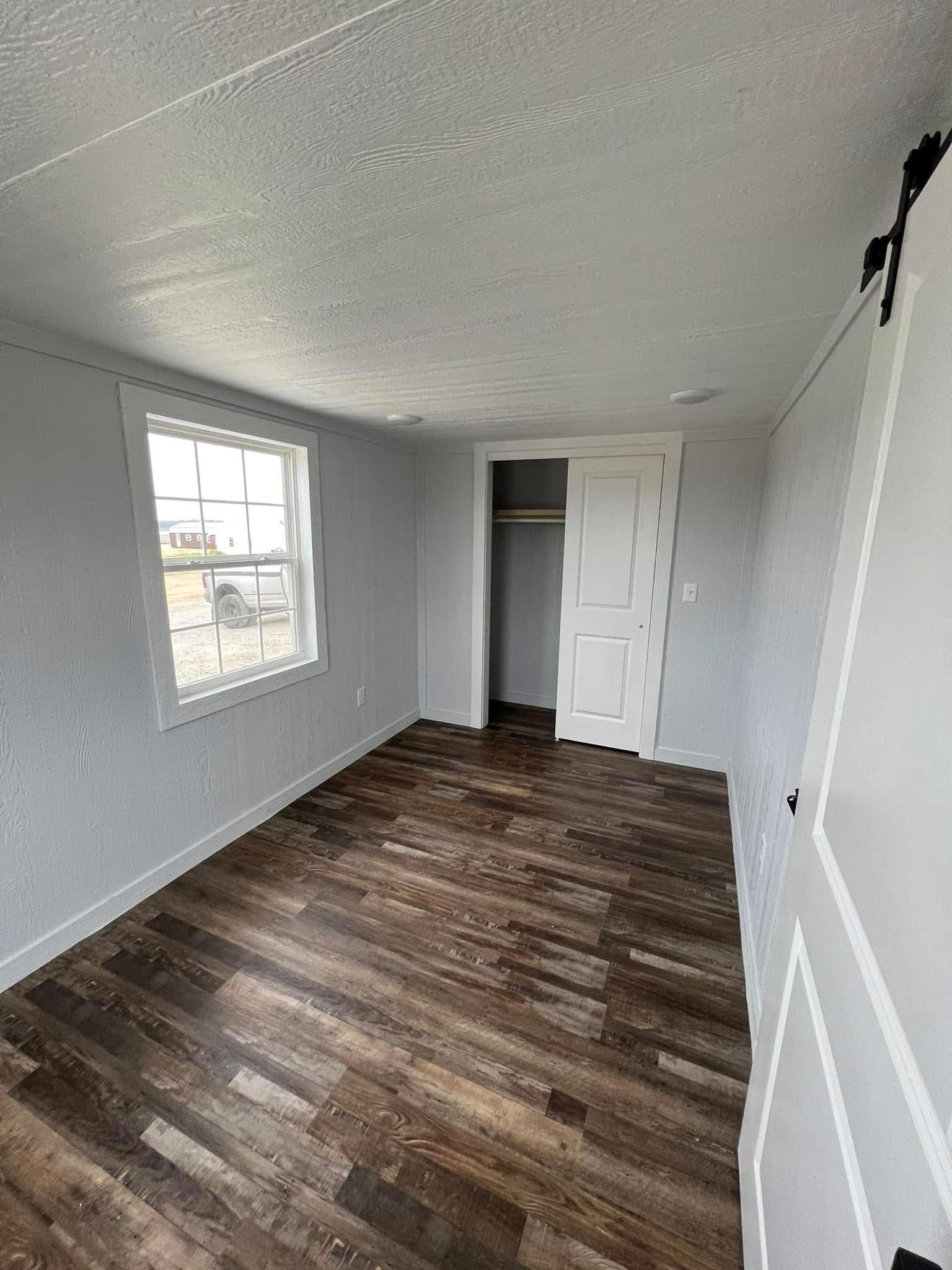 Small empty room with white walls and wood flooring. A window on the left brings light, revealing a closet with an open white door at the far end.