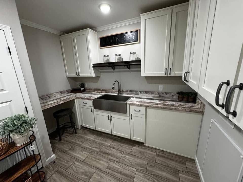 A modern laundry room with white cabinets, a metallic farmhouse sink, and marble countertops. Shelves display glass jars and a "Laundry" sign. The tone is sleek and functional.