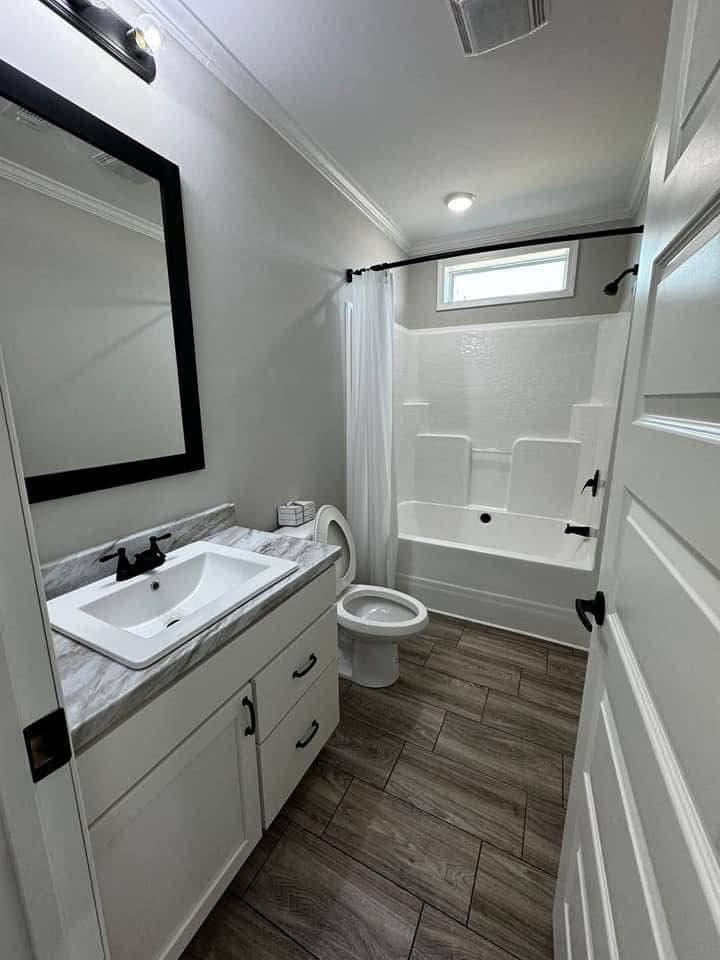 Compact modern bathroom with a white tub and shower, wooden floor tiles, marble vanity, black-framed mirror, and soft overhead lighting.