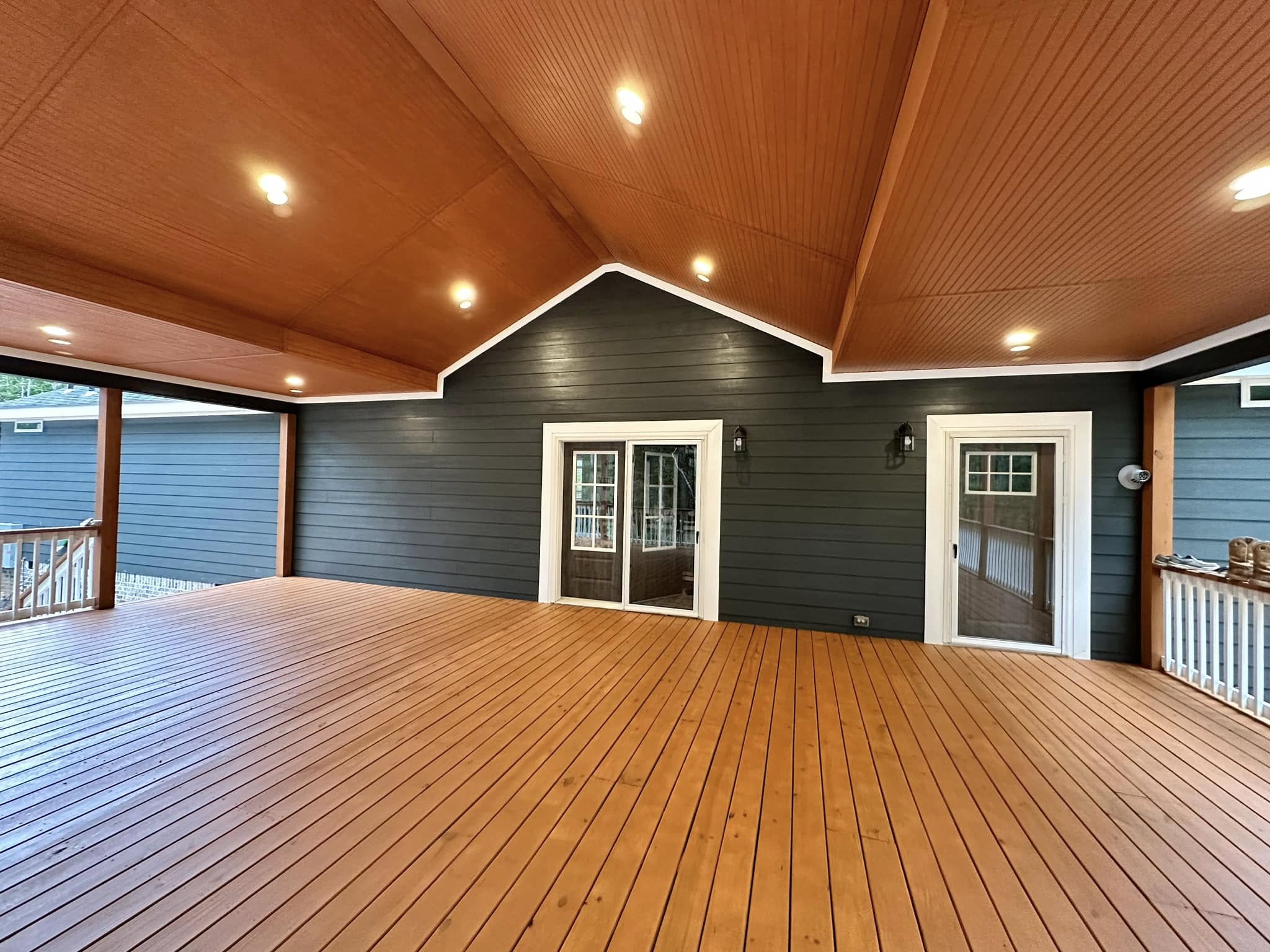 Covered wooden deck with recessed lighting and vaulted ceiling. Dark siding walls feature French doors, creating a cozy, inviting outdoor space.