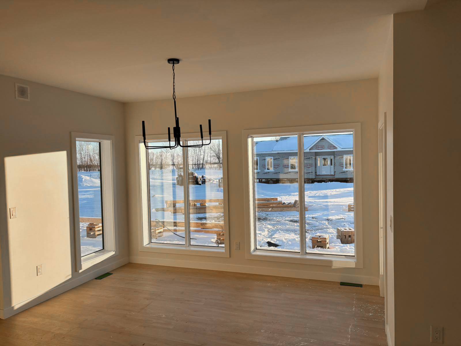 Minimalist room with large windows, natural light on wooden floors, and a modern black chandelier. Snowy landscape and distant house visible outside.