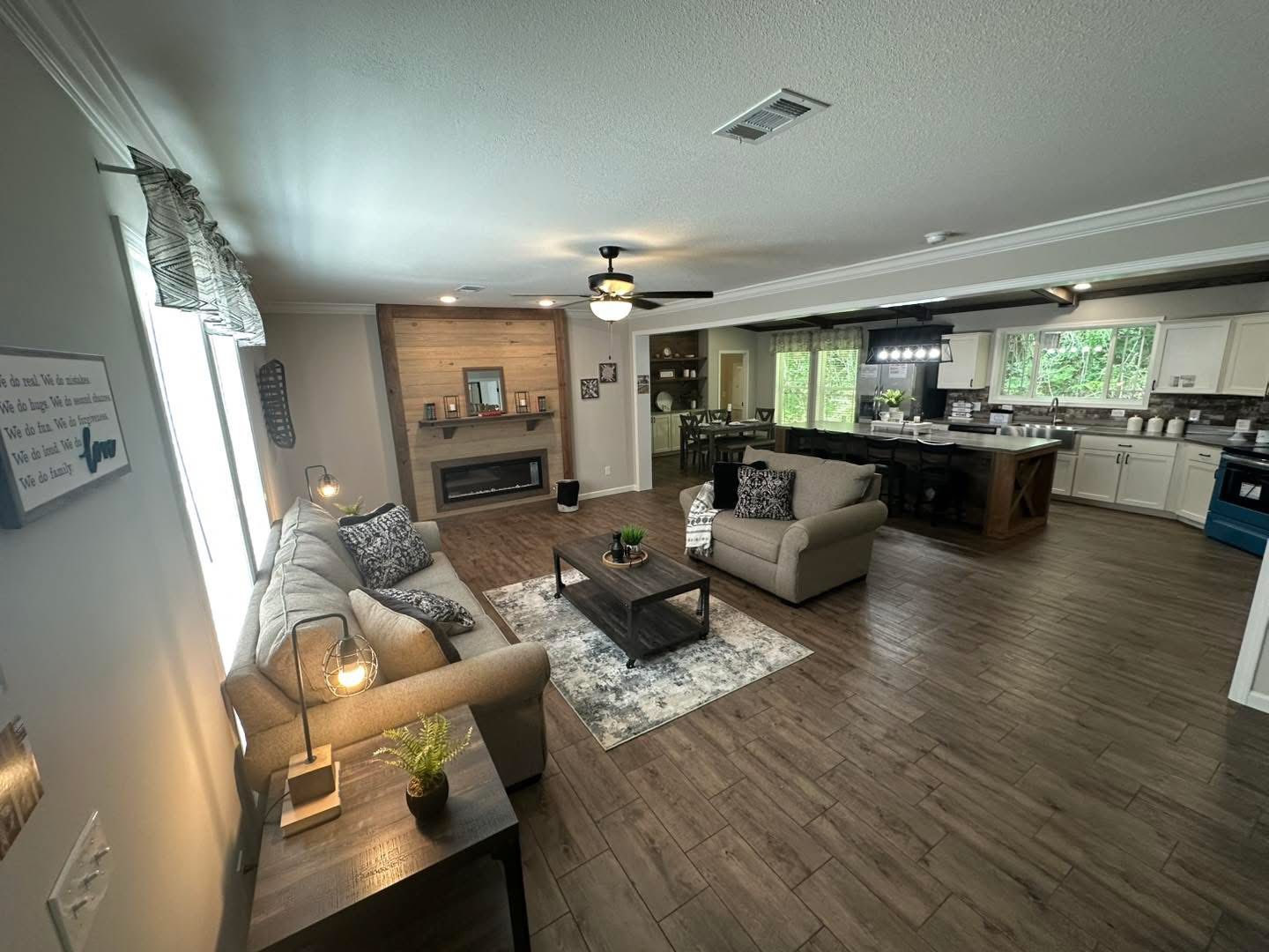 Spacious living room with a cozy seating area, warm wood accents, fireplace, and ceiling fan. Open layout extends to a modern kitchen with large windows.