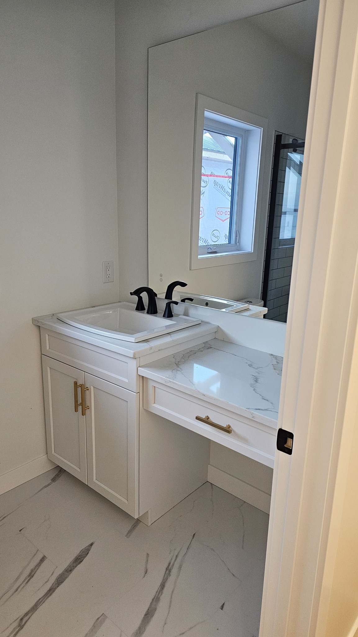 A modern bathroom corner with a white vanity featuring brass handles, a black faucet, and a large mirror. A window provides natural light. Neutral tones create a calm atmosphere.