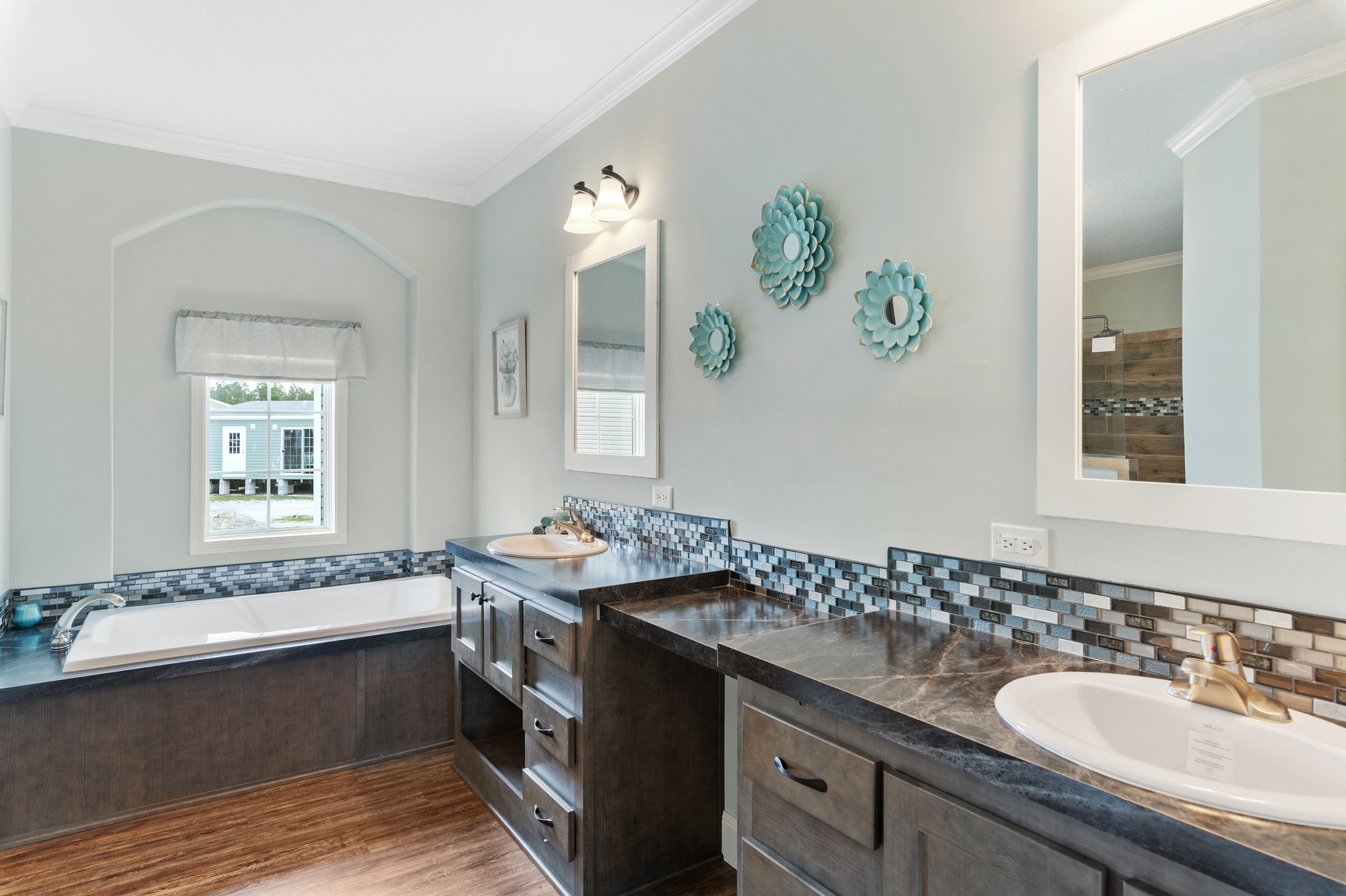 Stylish bathroom with double sinks on dark wood vanity, blue mosaic backsplash, and turquoise wall decor. Bathtub near a window adds a serene touch.