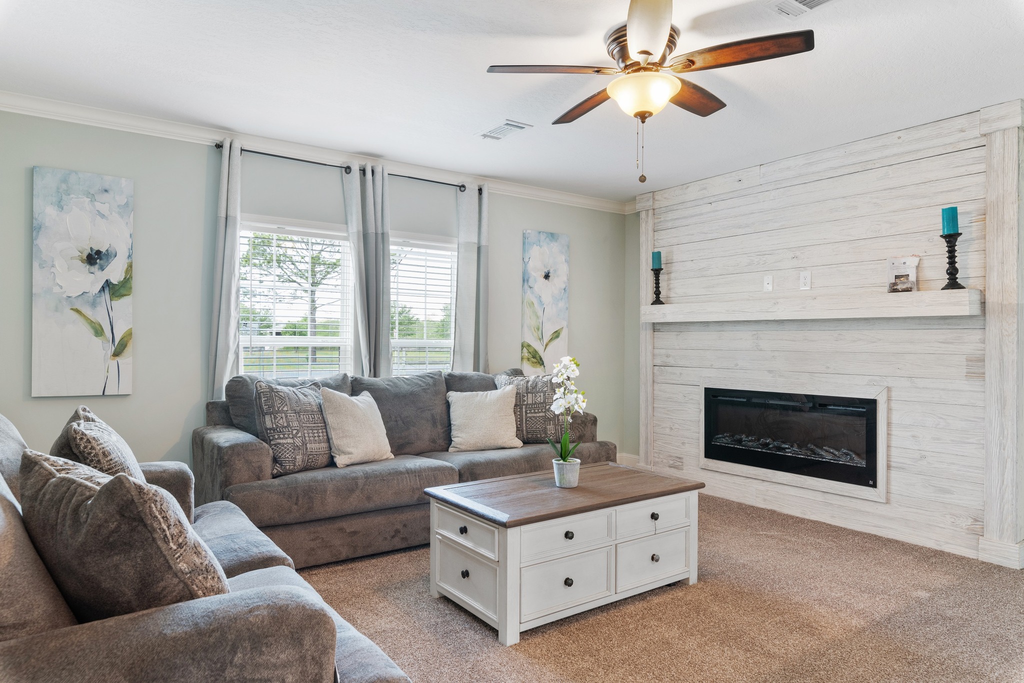 Cozy living room with gray sofas, floral cushions, and a light wood coffee table. Features a white shiplap fireplace, flower art, and gentle lighting.