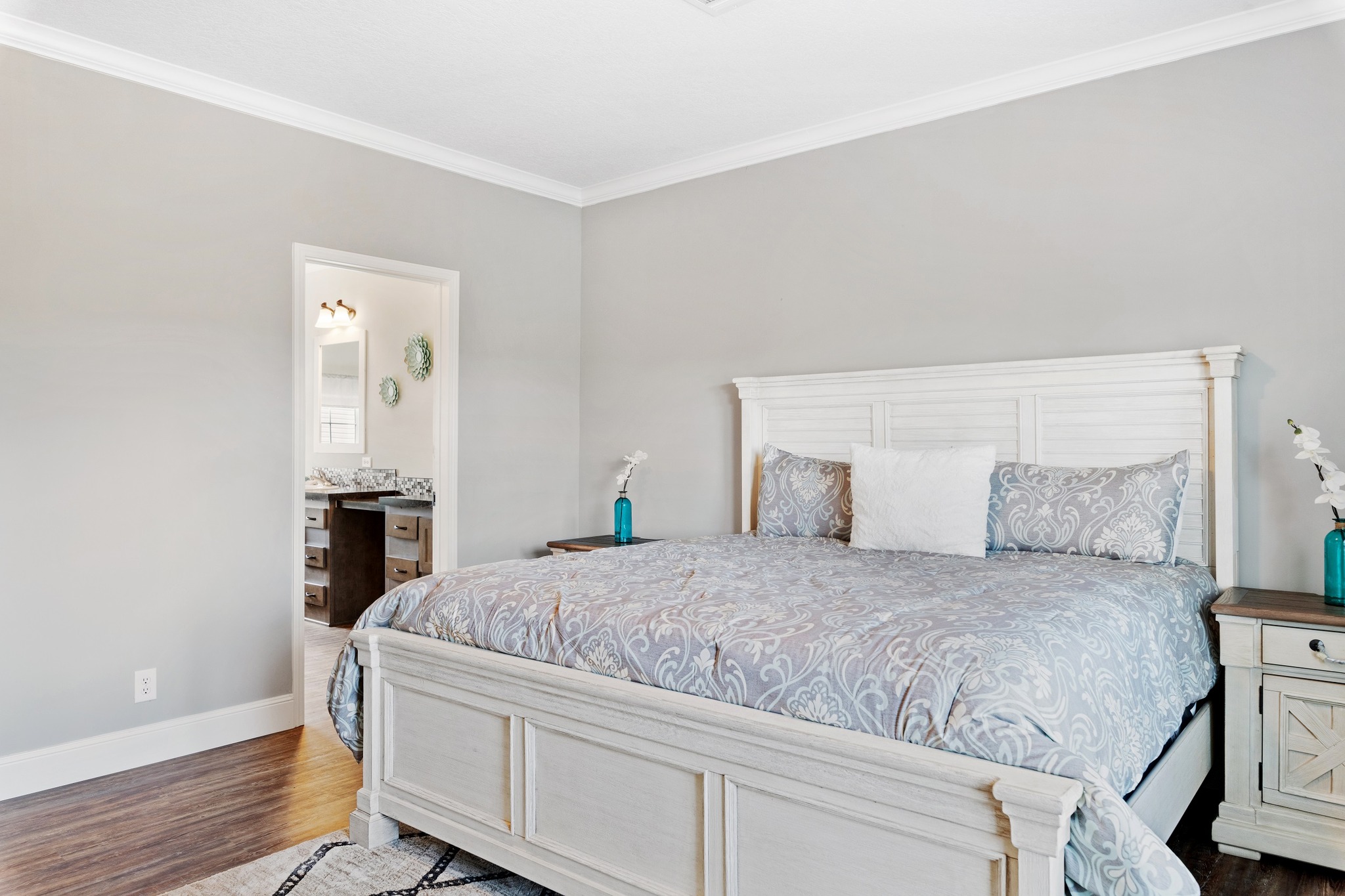 Bright, elegant bedroom with a large, ornate bed and matching nightstands. A doorway leads to a bathroom. The tone is calm and airy.