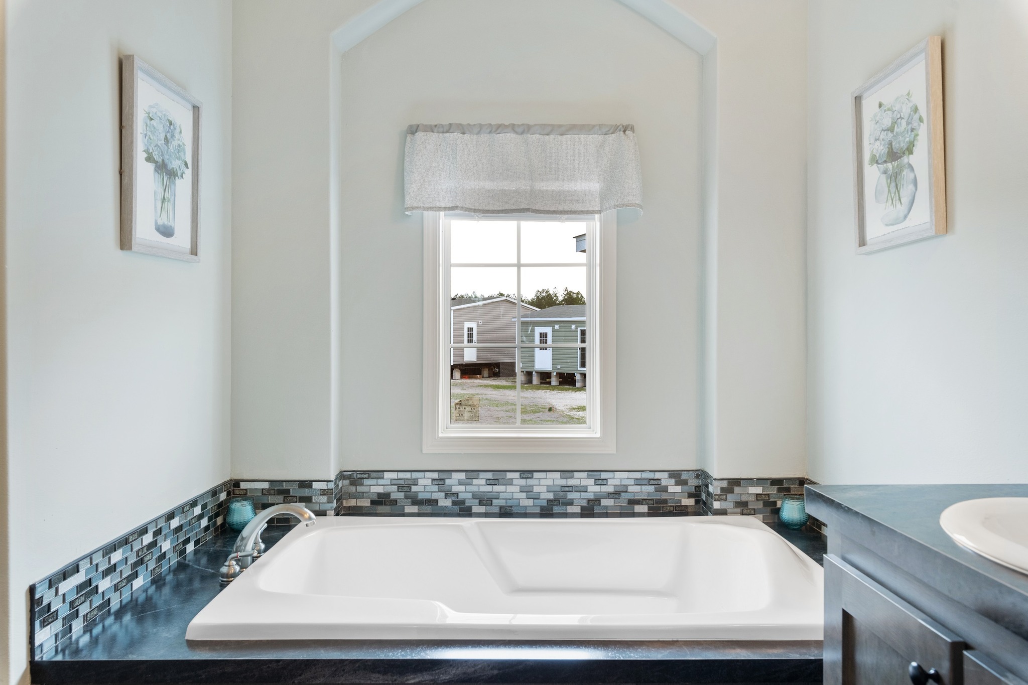 Bright bathroom with a white tub bordered by blue mosaic tiles. A window above overlooks houses. Floral paintings on light walls add elegance.