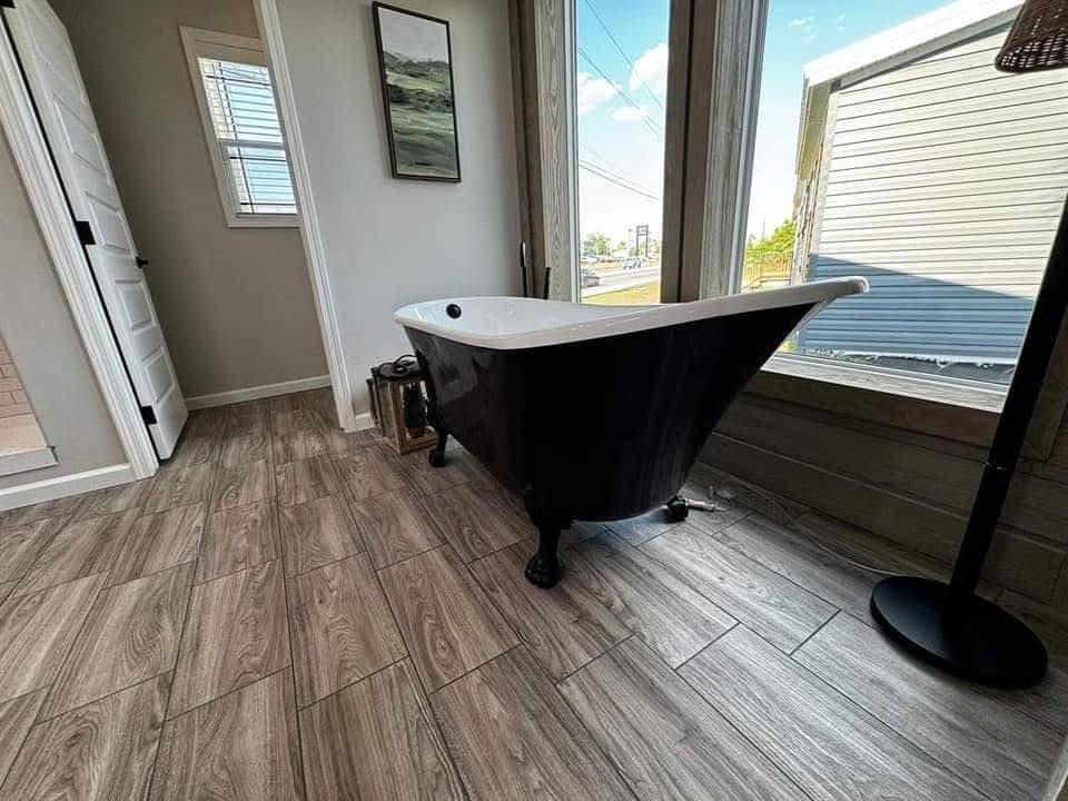Freestanding black bathtub in a sunlit room with wood flooring, large windows, and a serene outdoor view. Modern, calming atmosphere.