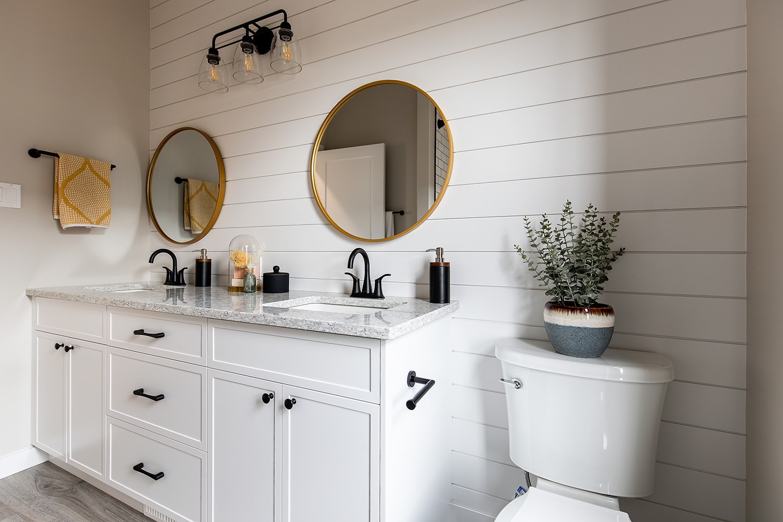 Modern bathroom with white shiplap walls, dual sinks on a white vanity with granite top, gold round mirrors, black fixtures, and a plant by a toilet.