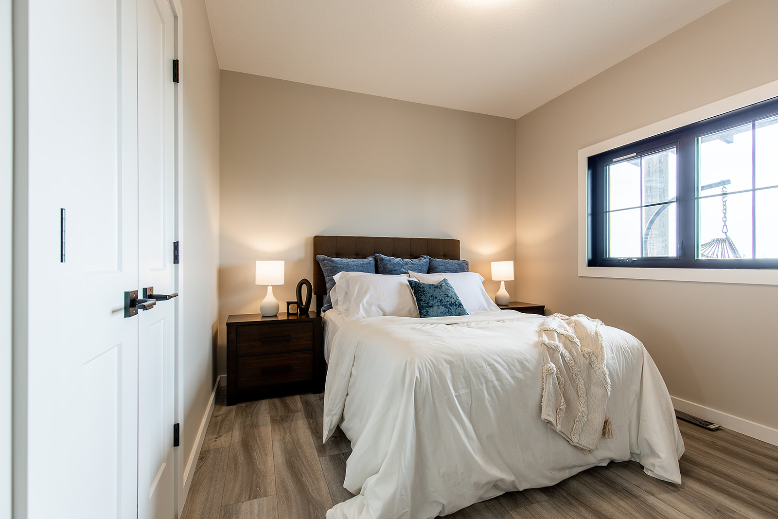 A cozy bedroom with a neatly made bed featuring white and blue pillows, flanked by wooden nightstands with lit lamps, and a window letting in daylight.