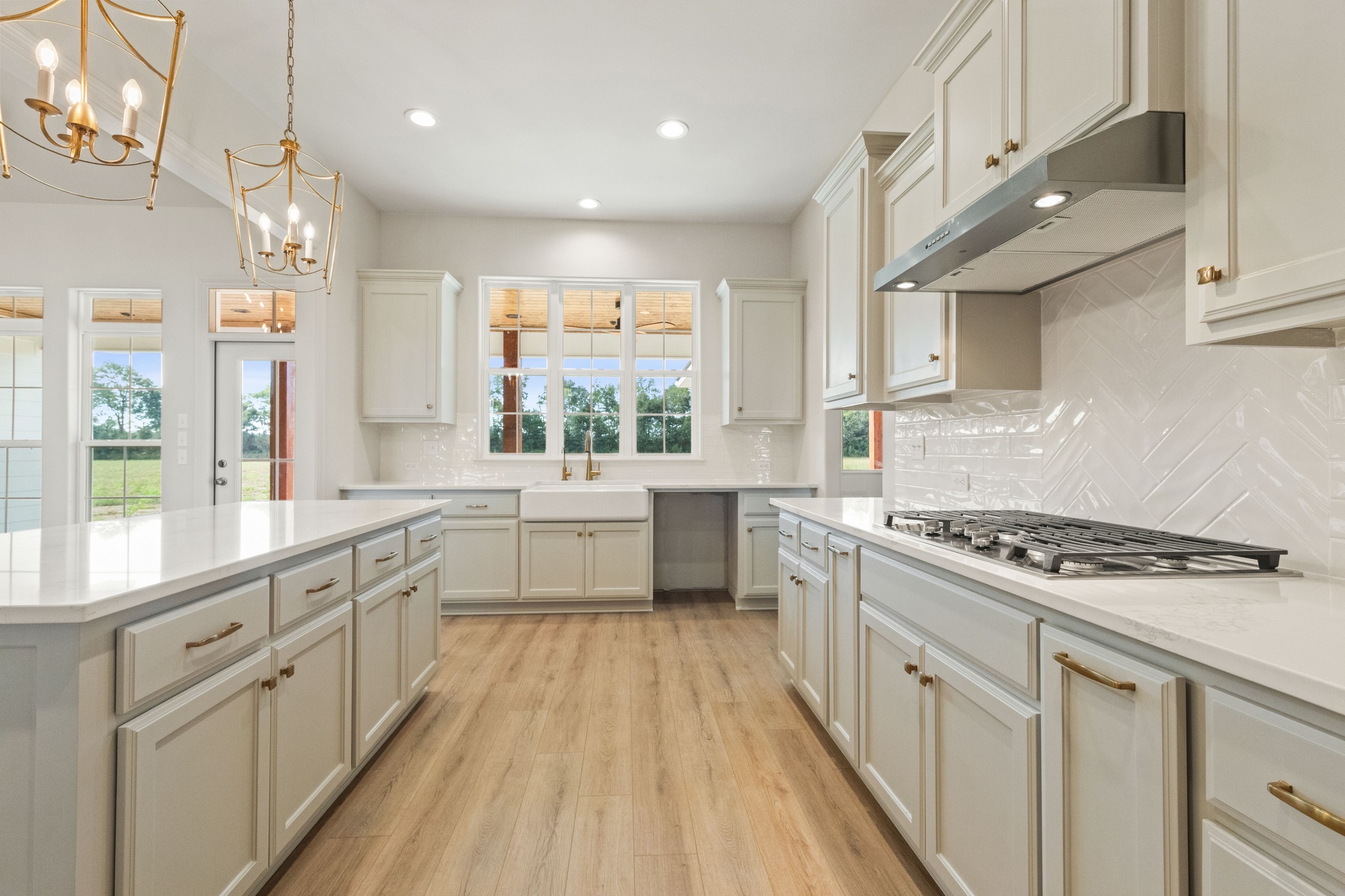 Spacious, elegant kitchen with white cabinets, hardwood floor, large island, stainless steel range, gold chandeliers, and bright windows overlooking greenery.
