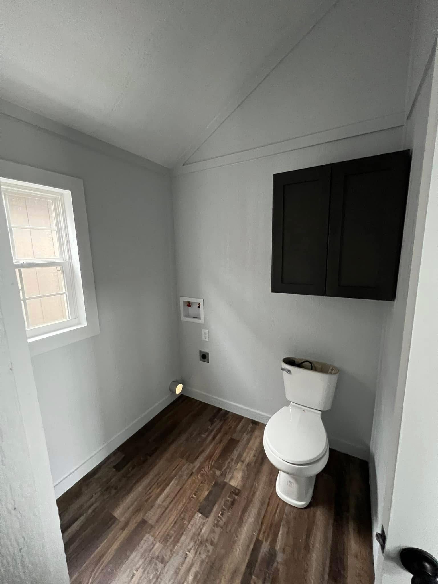 A minimalist bathroom with white walls features a single window, a wooden floor, a white toilet, and a dark-colored wall cabinet, creating a clean, modern look.