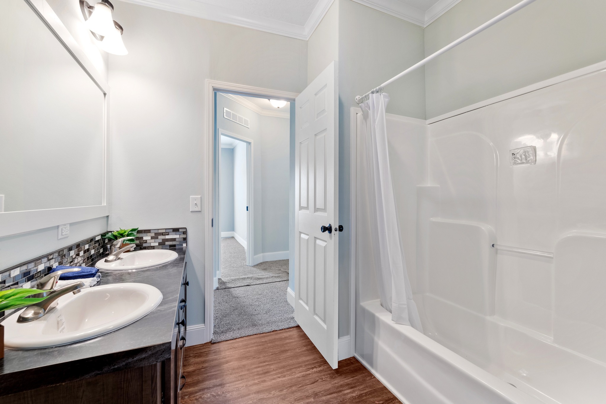 Bright, modern bathroom with dual sinks, mosaic backsplash, and a white tub with curtain. Open door leads to a carpeted hallway, evoking a clean, fresh feel.