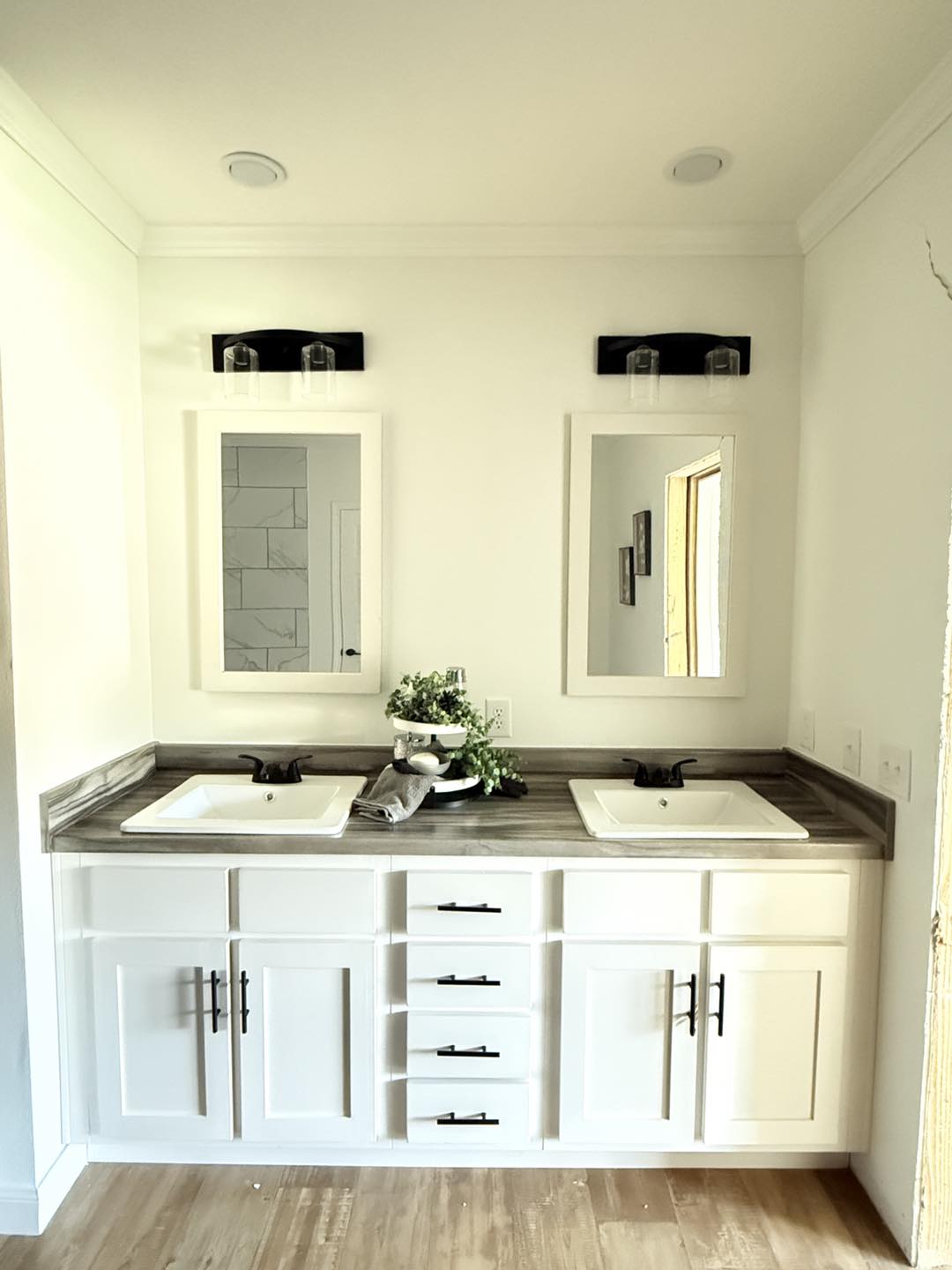 White bathroom vanity with two rectangular mirrors above dual sinks on a gray countertop. Black fixtures, white cabinets, and a small plant add a fresh, modern touch.