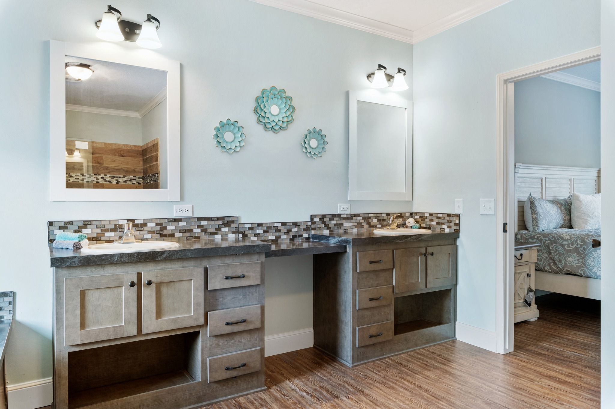 Modern bathroom with two mirrors, dual sinks, and wood cabinetry. Light blue walls feature decorative floral art. A doorway reveals a cozy bedroom.
