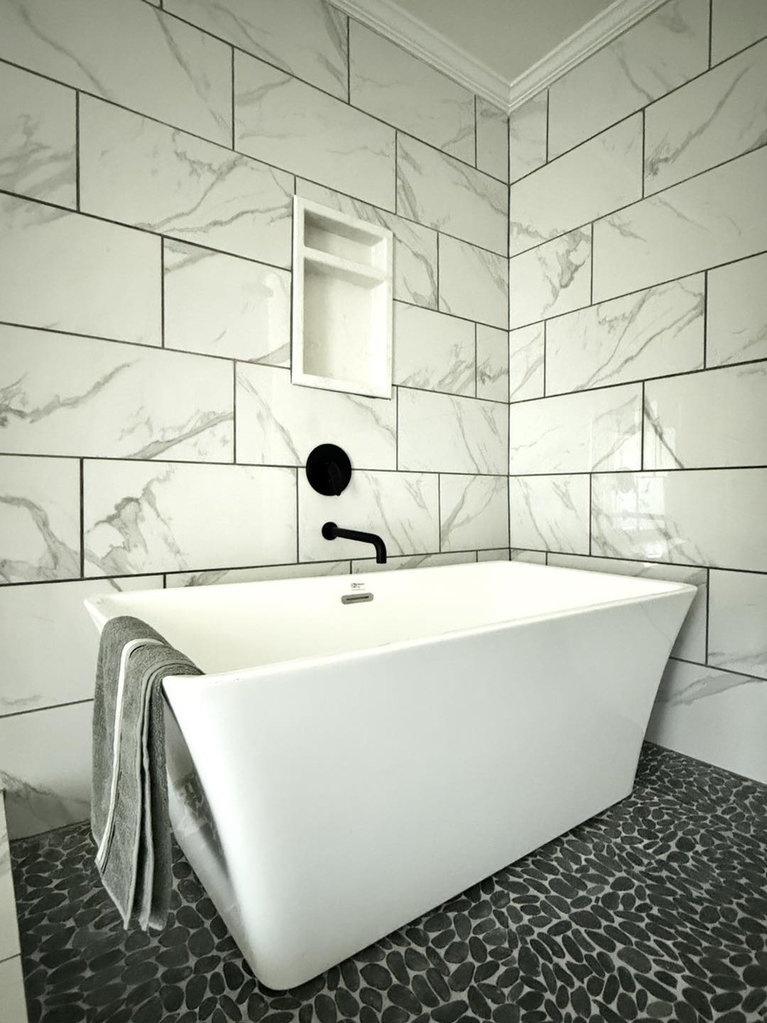 Modern bathroom with a white freestanding tub against gray marble tiled walls. Black fixtures and pebble flooring add contrast. A gray towel hangs over the tub edge.