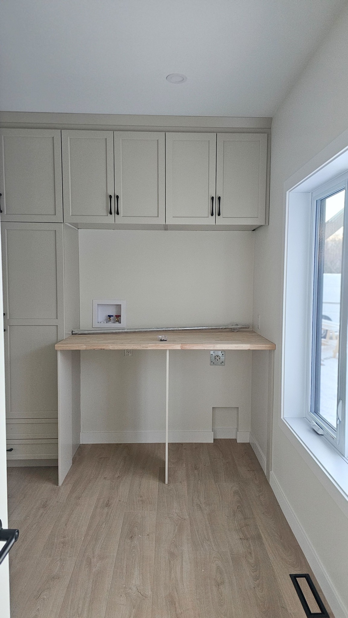 Minimalist light-toned workspace with a built-in wooden desk, white cabinets above, and a large window on the right. The room feels bright and airy.
