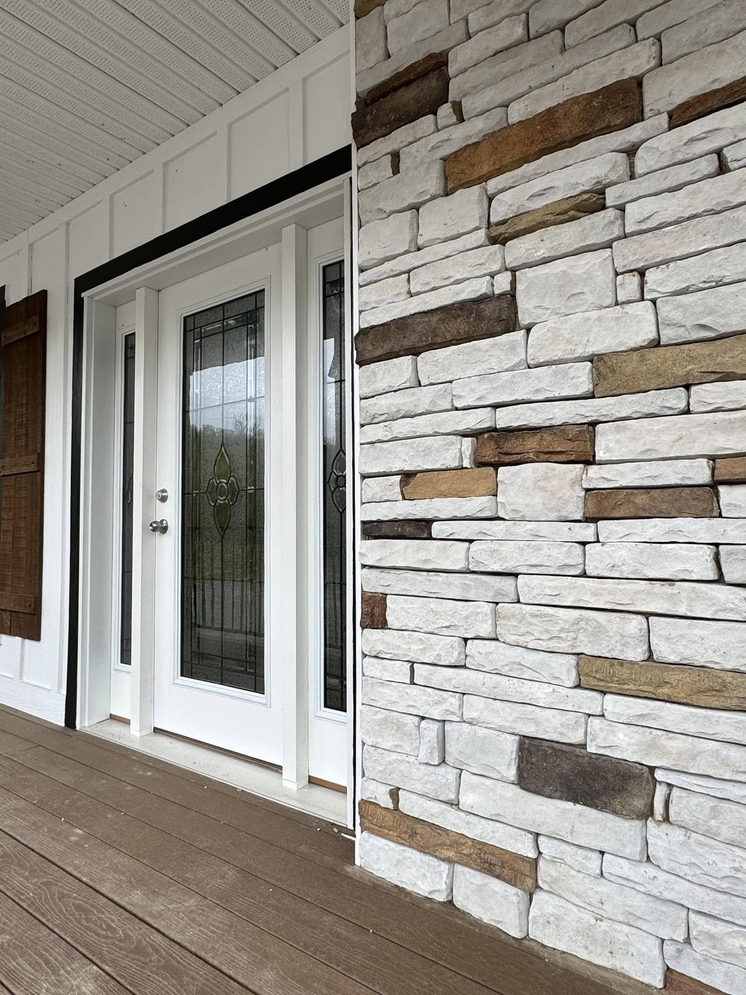 White double doors with decorative glass panels are set next to a wall of stacked stone in earthy tones. The setting suggests modern home entrance.
