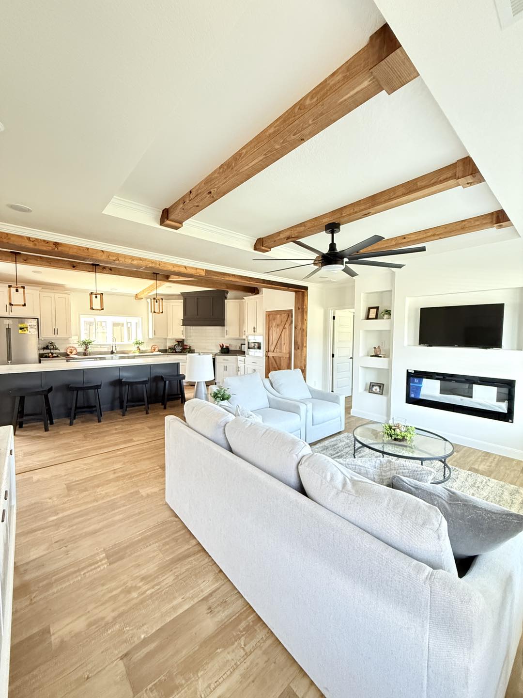 Modern living area with white furnishings, wooden beams, and a ceiling fan. Open kitchen with island and bar stools. Bright, inviting space.