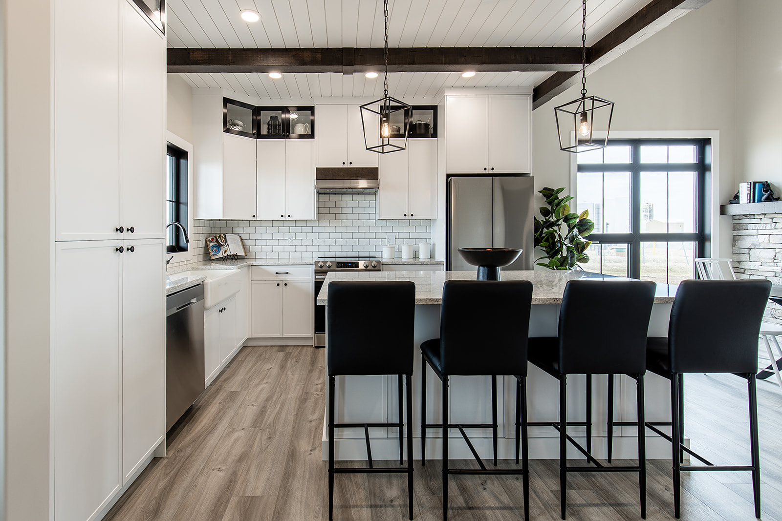 Stylish modern kitchen, featuring white cabinets, black bar stools, island with pendant lights, stainless steel appliances, and a large window.