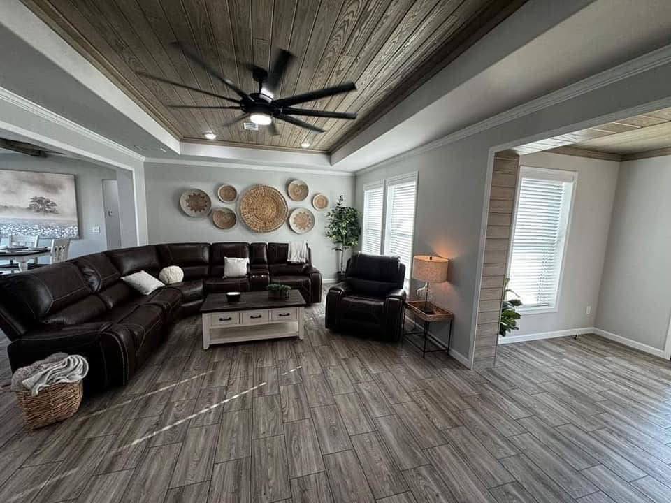 Spacious, cozy living room with dark leather sectional, recliners, and textured wall art. Wood ceiling, large fan, and abundant natural light.