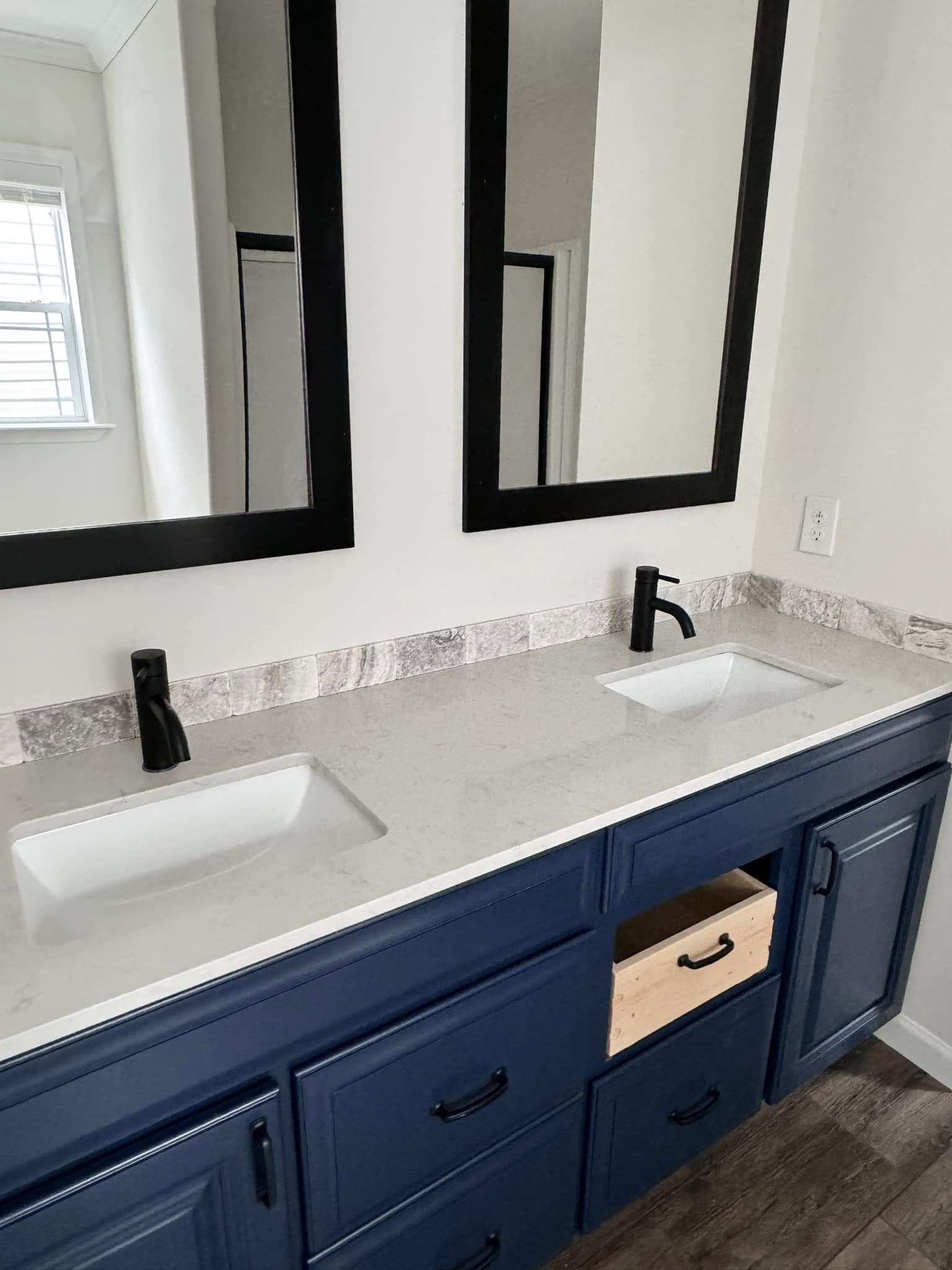 Blue bathroom vanity with dual sinks and black fixtures. Two large mirrors with black frames hang above. One drawer is slightly open. Bright ambiance.