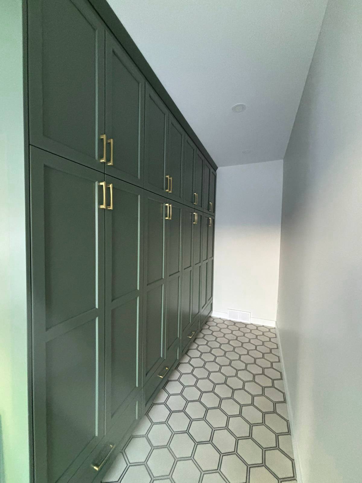 A narrow hallway with dark green cabinets featuring gold handles on the left, and a light hexagonal tile floor. The white walls create a modern, clean look.