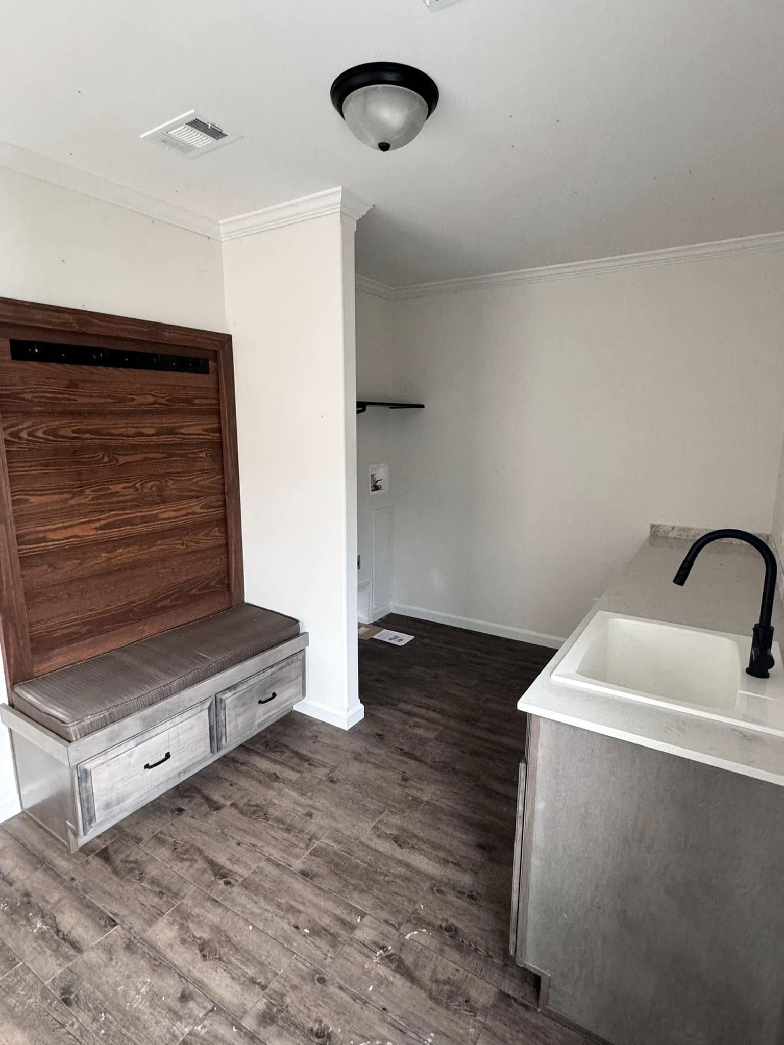 Small utility room with a wooden bench and coat hooks on the left, white sink with a black faucet on the right, and wooden floors throughout.