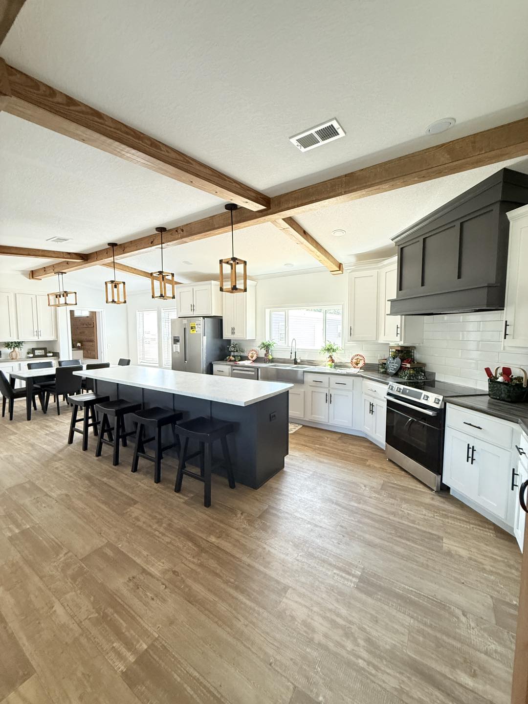 Spacious modern kitchen with wooden beams, white cabinets, a large island with black stools, and pendant lights. The space is bright and inviting.